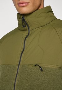 Zelená prešitá bunda s vysokým golierom, so zapínaním na zips a vreckom na hrudi, textúrovaná látka a hladká dolná časť.