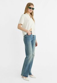 Camicia bianca a maniche corte con bottoni, jeans blu a vita alta e décolleté bianchi a punta; accessoriate con occhiali da sole verdi a forma rettangolare.