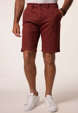 Mand iført opkrogede vinrøde shorts, hvide sneakers og en hvid skjorte, stående med den ene hånd i lommen og den anden ved siden.