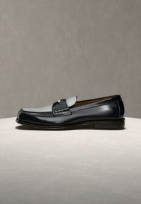 Μαύρο και γκρι δερμάτινο μοκασίνι με ασημένια αγκράφα, εμφανιζόμενο σε προφίλ πάνω σε επιφάνεια με ελαφρύ υφή και γκρι φόντο.