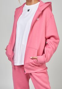Roze zip-up hoodie van zachte stof met een trekkoord capuchon, voorzakken en geborduurd logo, gecombineerd met een witte T-shirt en bijpassende broek.