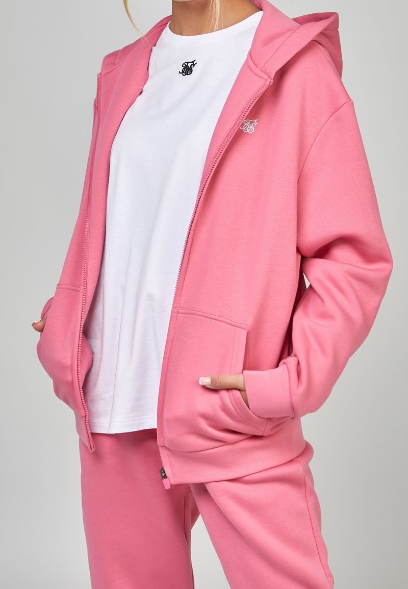 Felpa rosa con zip realizzata in tessuto morbido, con cappuccio regolabile, tasche frontali e logo ricamato, abbinata a una maglietta bianca e pantaloni coordinati.