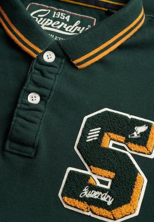 Mørkegrøn polo med en struktureret krave, der har orange striber. Den har en hvid knapstolpe og en broderet "S"-logo med accenter.