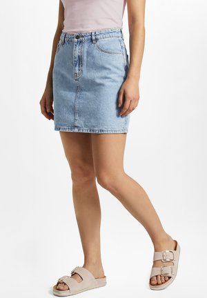 Frau trägt hellblauen Jeans-Minirock, blassrosa Oberteil und beige Slip-On-Sandalen mit zwei Schnallen, steht mit einem Bein nach vorne.