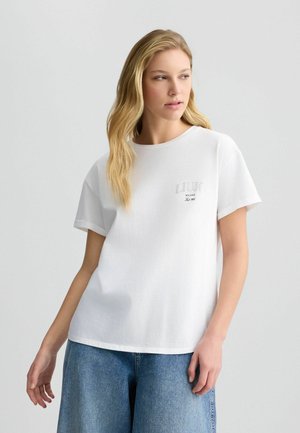 T-shirt bianca in cotone con maniche corte, scollo rotondo e un logo grigio sul petto. Presenta una vestibilità rilassata e una texture morbida.