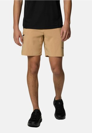 Homme portant un short beige avec une poche latérale et des chaussures de sport noires, debout devant un fond clair uni.