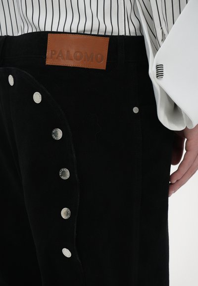 Pantaloni neri con chiusure a scatto laterali, dotati di un'etichetta in pelle "PALOMO" sulla cintura e una texture del tessuto liscia.