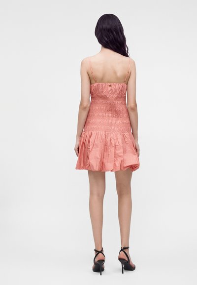 Femme portant une robe mini rose froncée sans manches et des sandales noires à talons hauts, debout dos à la caméra.