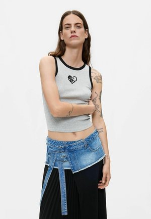 Vrouw met lang bruin haar die een grijze mouwloze top draagt met een zwart hartontwerp en een blauwe spijkerbroeksriem over een zwarte rok.