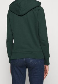 Felpa verde scuro con cappuccio regolabile, polsini e orlo a costine. Tessuto morbido, design semplice e cuciture decorative visibili sul retro.