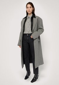 Manteau en laine gris avec un col en cuir noir, superposé sur une chemise à rayures noir et blanc, associé à un jean foncé et des bottines noires à bout pointu.