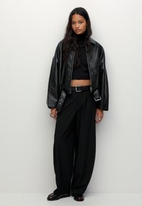 Črna usnjena bomber jakna s sproščenim krojem, skrajšano dolžino in stranskimi žepi, nosi se čez črn pulover z visokim ovratnikom in široke hlače.