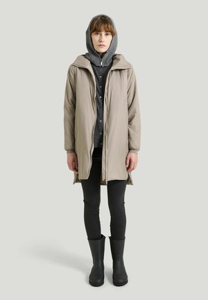 Ung person iført en beige frakke over en grå hættetrøje, mørke leggings og sorte gummistøvler mod en ensfarvet baggrund.