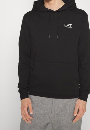 Mann trägt einen schwarzen EA7 Emporio Armani Hoodie mit Fronttasche und Kordeln, kombiniert mit hellgrauen Jogginghosen.