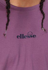 Fialové bavlnené tričko s okrúhlym výstrihom a čiernym logom "ellesse" na prednej strane. Textília má hladkú štruktúru.