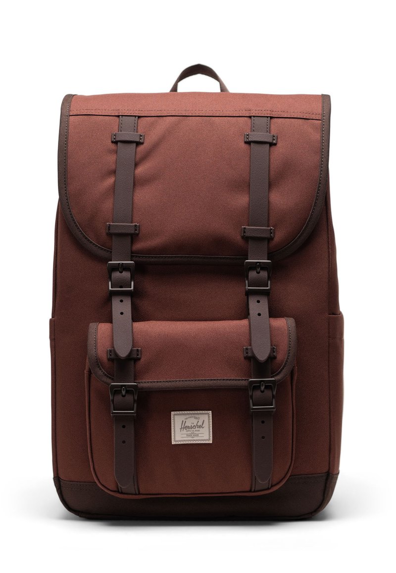 Brown canvas backpack with a flap, front pocket, and brown accents. Features adjustable straps and a textured finish.
