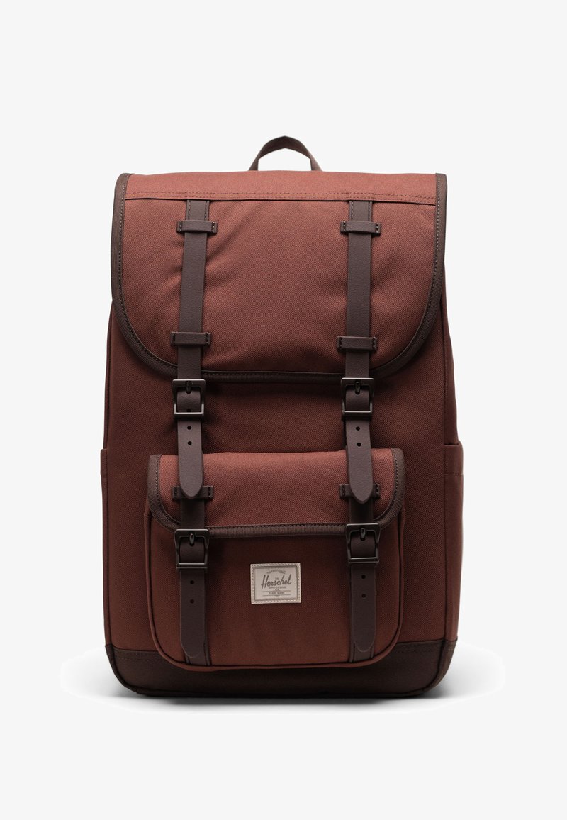 Brown canvas backpack with a flap, front pocket, and brown accents. Features adjustable straps and a textured finish.