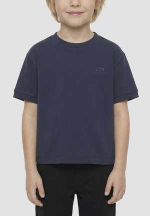 Jeune enfant aux cheveux clairs portant un t-shirt bleu marine uni et un pantalon noir, debout sur un fond neutre.
