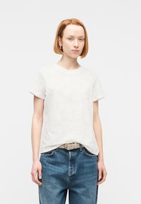 Νεαρός με κοντά κόκκινα μαλλιά που φοράει λευκό μπλουζάκι με λουλουδένιο μοτίβο, μπλε τζιν και μπεζ ζώνη, στέκεται μπροστά σε απλό φόντο.