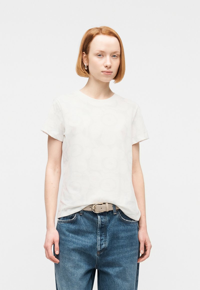 Νεαρός με κοντά κόκκινα μαλλιά που φοράει λευκό μπλουζάκι με λουλουδένιο μοτίβο, μπλε τζιν και μπεζ ζώνη, στέκεται μπροστά σε απλό φόντο.