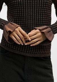 Sort tekstureret sweater med et gittermønster, der har volanger ved håndleddene i en metallisk bronze nuancer. Tætsiddende design, der fremhæver hænderne der er foldet sammen.