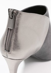 Stivale alla caviglia grigio con finitura in pelle metallica, tacco a stiletto e una prominente zip laterale. Texture liscia dal design elegante.