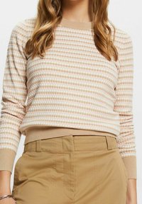 Vrouw draagt een beige en wit gebreid trui met een patroon, voorzien van geribde mouwen en onderkant, gecombineerd met lichtbruine hoge taille broek.