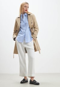 Manteau trench beige, chemise boutonnière bleu clair, pantalon large blanc et mocassins noirs. Textures lisses, ajustement sur mesure, palette de couleurs neutres.