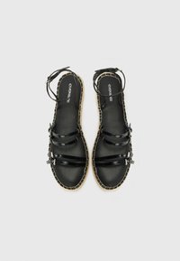 Sandalias de charol negro con tres correas, un hebilla en el tobillo y una suela trenzada en tono marrón. Textura suave y diseño minimalista.