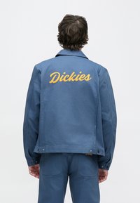 Μπλε εργασιακό μπουφάν με λείο ύφασμα, με κεντημένο λογότυπο "Dickies" σε κίτρινο χρώμα στην πλάτη και κουμπώματα με πατιλέτα στο τελείωμα.