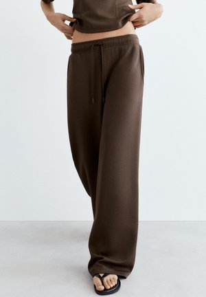 Person wearing loose brown sweatpants and black flip-flops, lifting their top slightly to reveal the waistband.