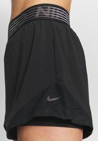 Mustat urheilushortsit, joissa on verkkokangaspaneeli, harmaa Nike-logo helmassa ja leveä joustava vyötärönauha, jossa on hopeanväriset raidat.