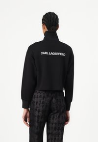 Čierna skrátená mikina s vysokým golierom a vyšitým logom "KARL LAGERFELD" na chrbte, v kombinácii so vzorovanými tmavými nohavicami.