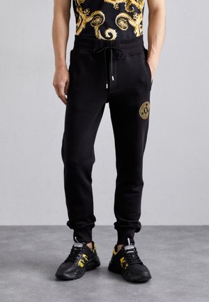 Person trägt schwarze Jogginghosen mit goldenem Logo am Oberschenkel, kombiniert mit schwarz-gold gemusterten Sneakers und einem passenden schwarz-goldenen Oberteil.