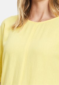 Żółta, lekka bluzka z okrągłym dekoltem, krótkimi rękawami i miękką fakturą; posiada detal szycia na przodzie.
