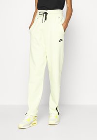 Nike joggers in pale yellow, featuring an elastic waistband with black drawstring and side pockets. Black logo on left thigh.