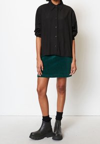 Schwarze Button-Down-Hemd mit Kragen, kombiniert mit einem grünen Cordrock und schwarzen klobigen Stiefeletten, mit gerippten Strümpfen.