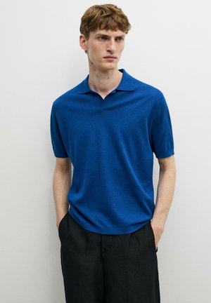 Giovane uomo che indossa una polo blu a maniche corte e pantaloni neri, in piedi con le mani in tasca su uno sfondo chiaro e semplice.