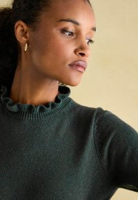 Dunkelgrüner Pullover mit gerüschtem Kragen und strukturiertem Stoff. Nahaufnahme, die das Kragenmuster und das weiche Material hervorhebt.