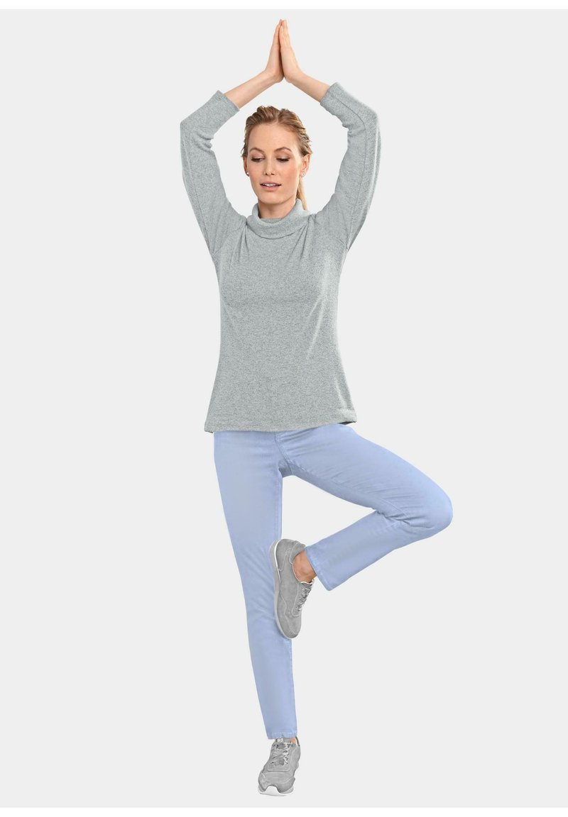 Grijze lange mouwen top met een col, gecombineerd met lichtblauwe broek. Het model staat in een yoga-houding, met grijze sportschoenen aan.