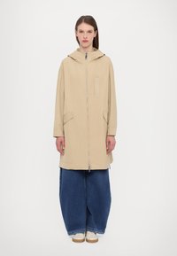 Giacca beige con cappuccio, chiusura frontale con zip, due tasche laterali e una toppa di tessuto. Abbinata a pantaloni in denim blu a gamba larga e scarpe chiare.