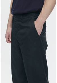 Black cotton trousers with a straight cut, featuring side pockets and a button closure at the waistband. Smooth texture, minimal design.