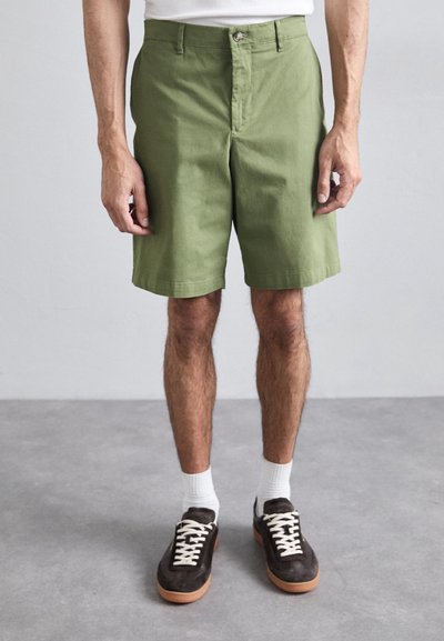Pantaloni corti verdi in cotone con vestibilità slim, dotati di chiusura a bottone e tasche laterali. Indossati con calze bianche e sneaker marroni.