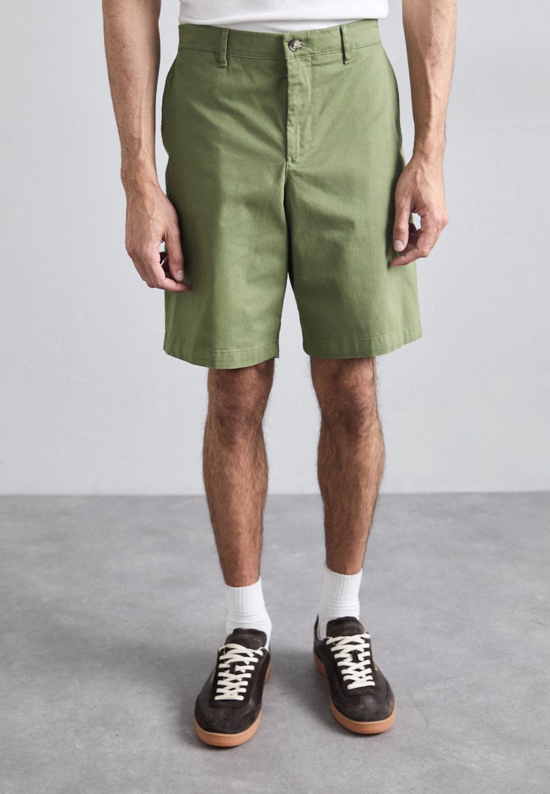 Shorts en coton verts à coupe slim, avec une fermeture à bouton et des poches latérales. Portés avec des chaussettes blanches et des baskets marron.