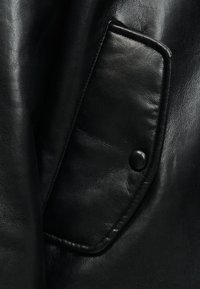 Poche de veste en cuir noir avec rabat et un seul bouton noir, montrant une texture lisse et des coutures discrètes.