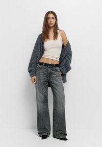 Szürke kötött kardigán egy bézs top fölött, széles szárú, fekete farmerral, amely csillaghímzéssel és fekete övvel van párosítva.