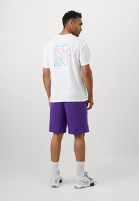 Hvid bomulds t-shirt med et blåt og pink grafisk "N" på ryggen, parret med lilla shorts. Har en afslappet pasform og rund halsudskæring.