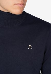 Maglione collo alto blu navy realizzato in materiale a costine, caratterizzato da un piccolo emblema ricamato in crema, che rappresenta oggetti incrociati con una forma di corona.