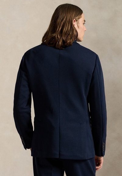 Man with shoulder-length brown hair wearing a dark navy suit jacket, facing away against a plain beige background.