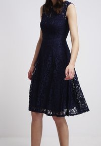 Navy blue lace dress with a fitted bodice and flared skirt. Floral pattern, sleeveless design, and knee-length hem. Textured lace fabric.
